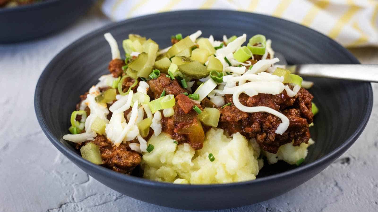 A bowl of Sloppy Joe mashed potatoes topped with chili, shredded cheese, chopped pickles, and green onions, with a spoon on the side.