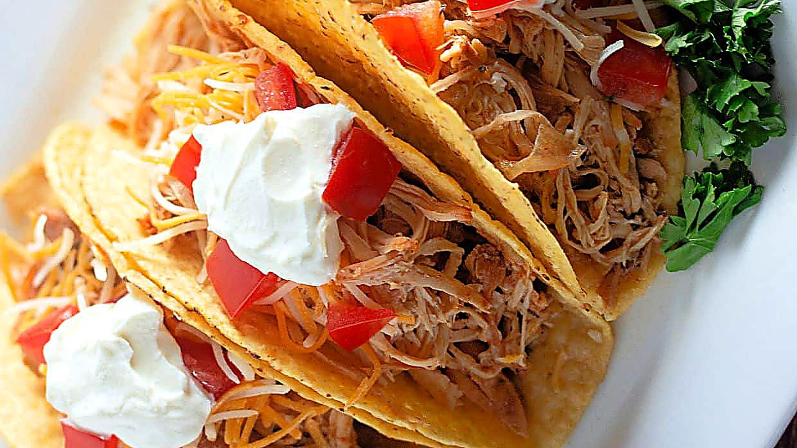 Three hard shell tacos filled with shredded slow-cooked chicken, cheese, diced tomatoes, and topped with sour cream, served on a white plate with a garnish of fresh parsley.