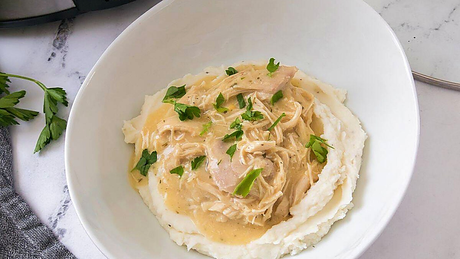 A bowl of mashed potatoes topped with shredded chicken in creamy gravy, garnished with chopped parsley.
