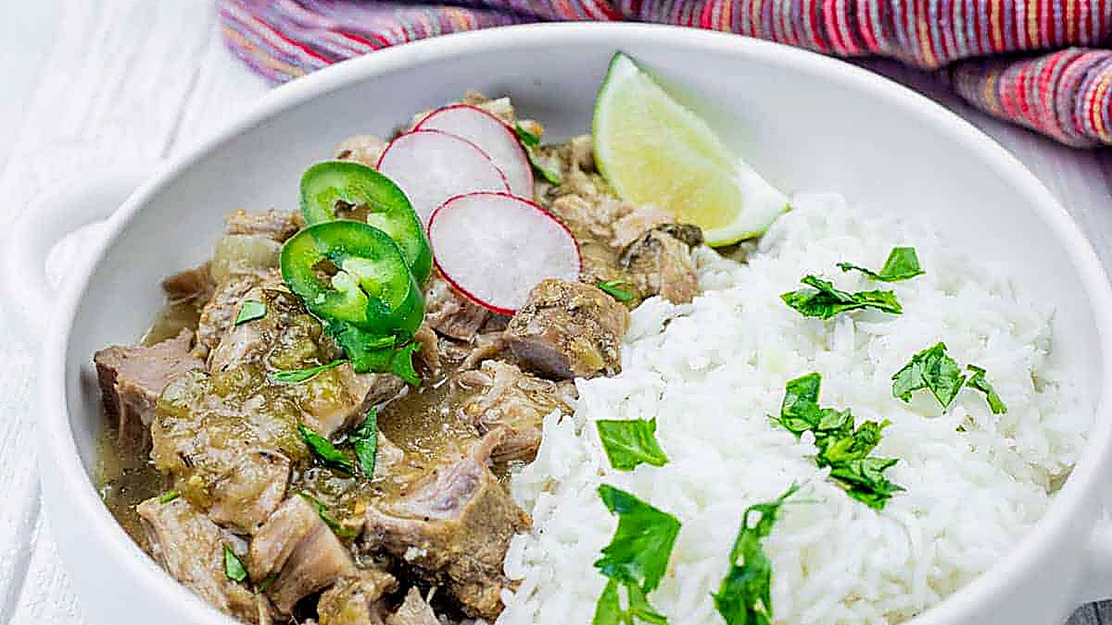 A bowl of white rice and green chili verde garnished with cilantro, radish slices, jalapeño, and a lime wedge, set on a white surface with a colorful cloth in the background.