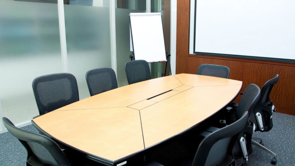 A conference room with eight black chairs around a wooden table, a blank flip chart, frosted glass panels, and a large white projection screen—ideal for those looking to get work done in airport environments or between flights.
