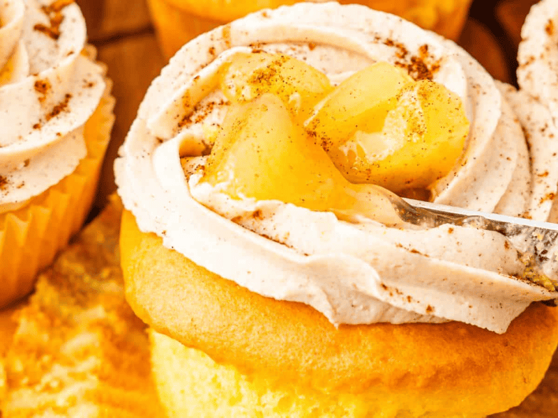 Close-up of cupcakes topped with frosting and apple chunks, sprinkled with cinnamon. A knife is spreading the frosting.