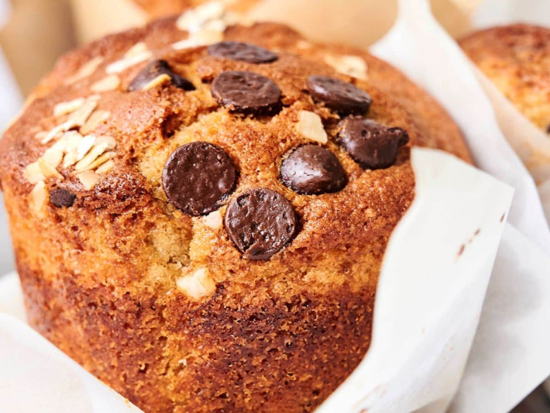 Close-up of a freshly baked muffin topped with chocolate chips and oats, wrapped in a paper liner. A blurred drink and other baked goods are visible in the background.