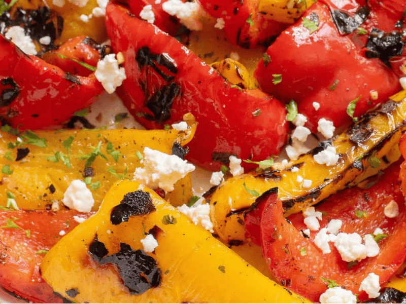 Grilled red and yellow bell peppers with char marks, topped with crumbled feta cheese and garnished with herbs.