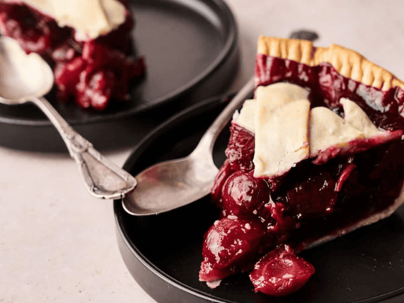 A slice of cherry pie with a flaky crust is served on a black plate with a spoon; another plate with pie is in the background.