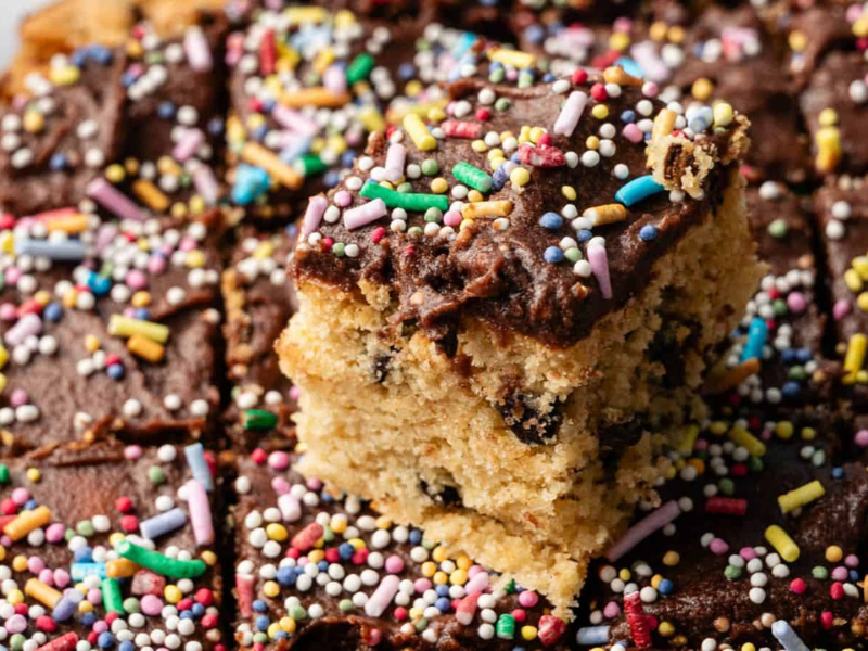 A tray of chocolate-frosted cake squares with colorful sprinkles, featuring one piece slightly raised above the rest.