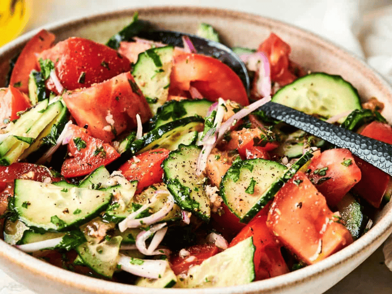 A bowl of fresh salad with sliced tomatoes, cucumbers, red onions, and herbs, seasoned with black pepper, with a serving spoon.