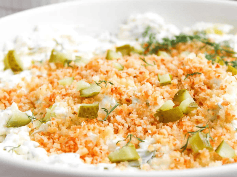 A close-up of a creamy dip topped with toasted breadcrumbs, chopped pickles, and fresh dill in a white bowl.