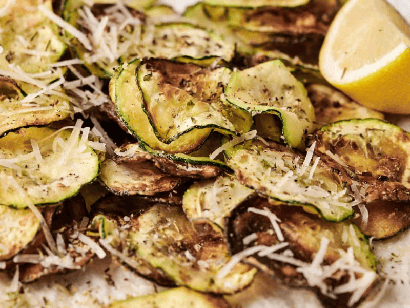 A plate of baked zucchini chips topped with shredded parmesan cheese and a lemon wedge on the side.