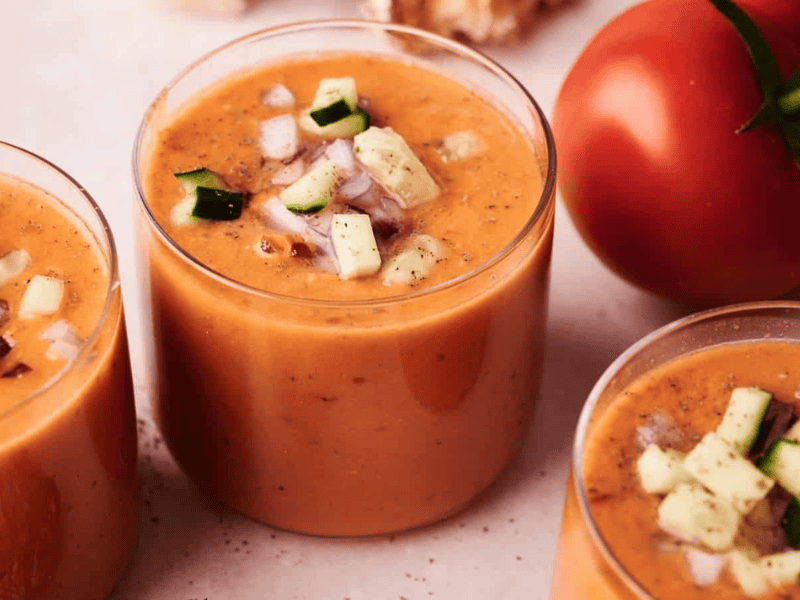 Three glasses of gazpacho soup topped with chopped cucumber and red onion, with a whole tomato in the background on a light surface.