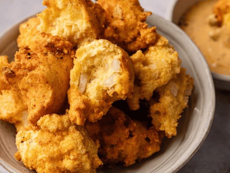 A bowl filled with golden, crispy hush puppies, some with visible onions, sits next to a small dish of dipping sauce.