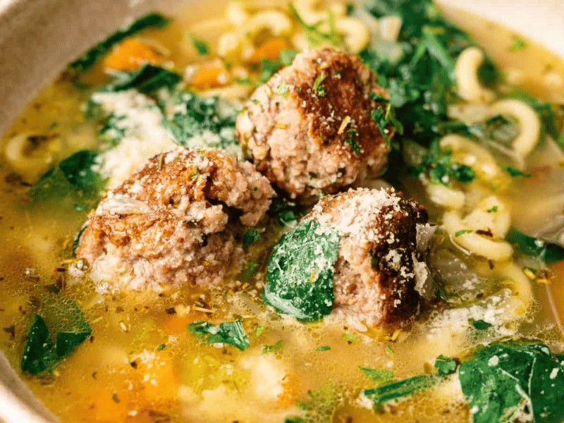 Bowl of soup with meatballs, spinach, pasta, and vegetables in broth, topped with grated cheese and herbs.