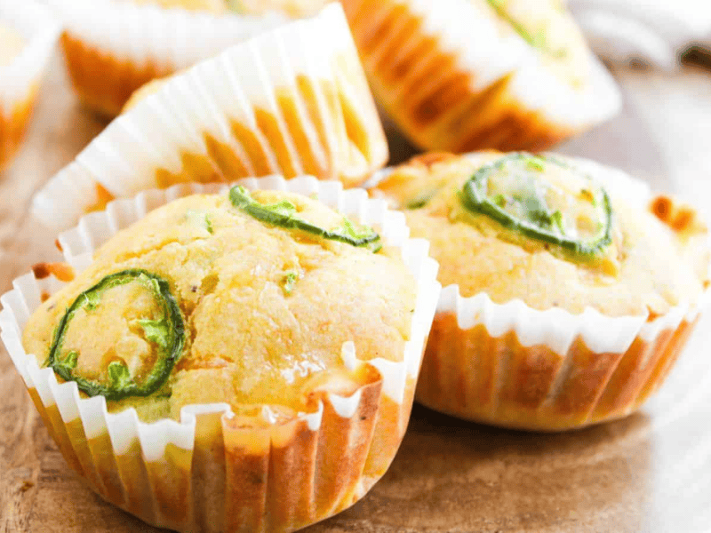 Cornbread muffins topped with sliced jalapeños in white paper liners, arranged on a wooden surface.