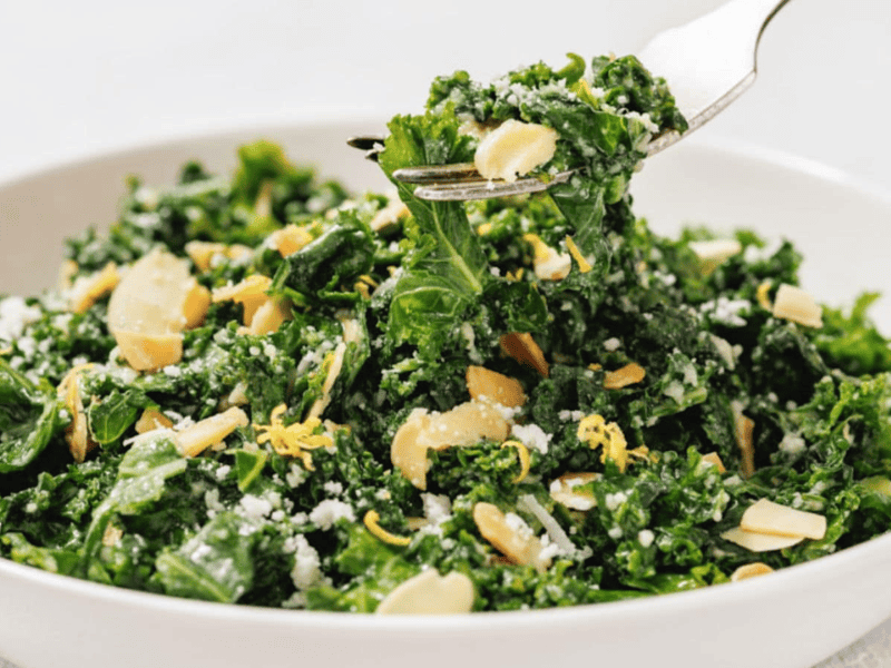 A bowl of kale salad topped with sliced almonds, grated cheese, and lemon zest, with a fork lifting a portion.