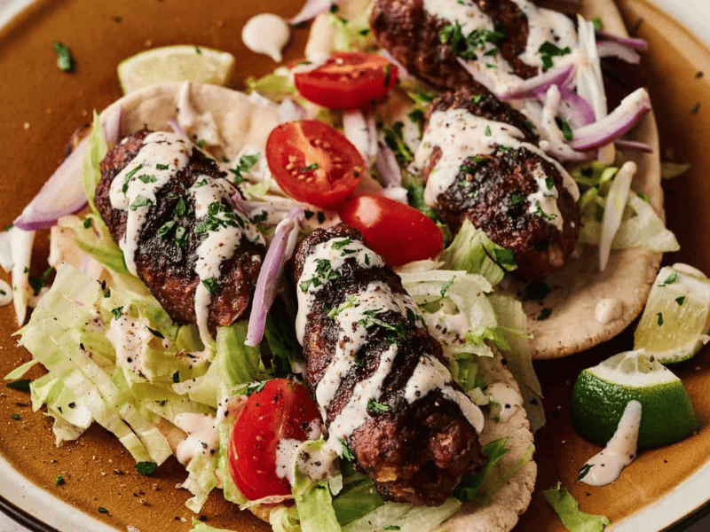 Three grilled kebabs on flatbread, topped with shredded lettuce, cherry tomatoes, red onion, chopped herbs, and a drizzle of white sauce, served with lime wedges.