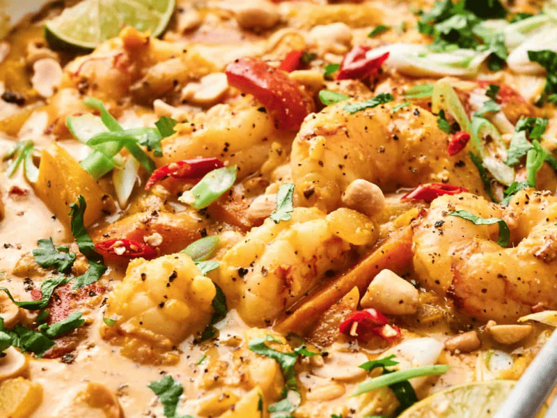 A creamy shrimp curry with peppers, chopped nuts, sliced green onions, red chili slices, and lime wedges. Metal spoon submerged in the dish.