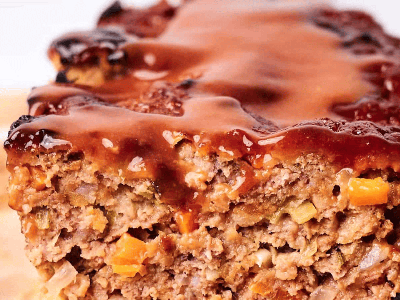 Close-up of a slice of meatloaf with visible chopped vegetables and a shiny layer of tomato glaze on top.