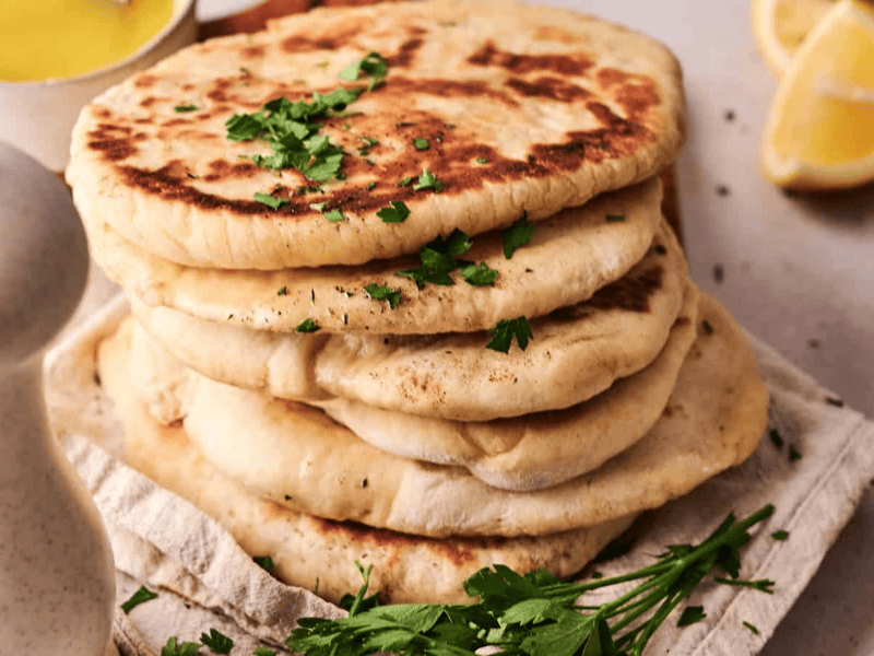 A stack of flatbreads garnished with chopped parsley sits on a cloth, with a white pepper grinder, fresh herbs, a lemon wedge, and a bowl of yellow sauce nearby.