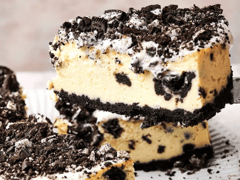 A slice of cookies and cream cheesecake is being lifted from a plate. The cake has a crumbly cookie crust, creamy filling with cookie pieces, and a topping of crushed cookies.