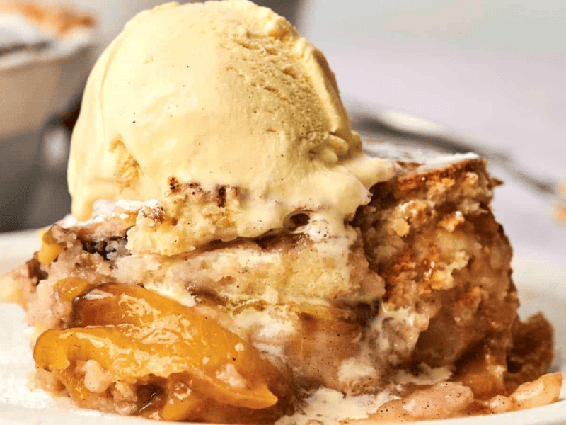 A close-up of a slice of peach cobbler topped with a scoop of vanilla ice cream on a white plate.