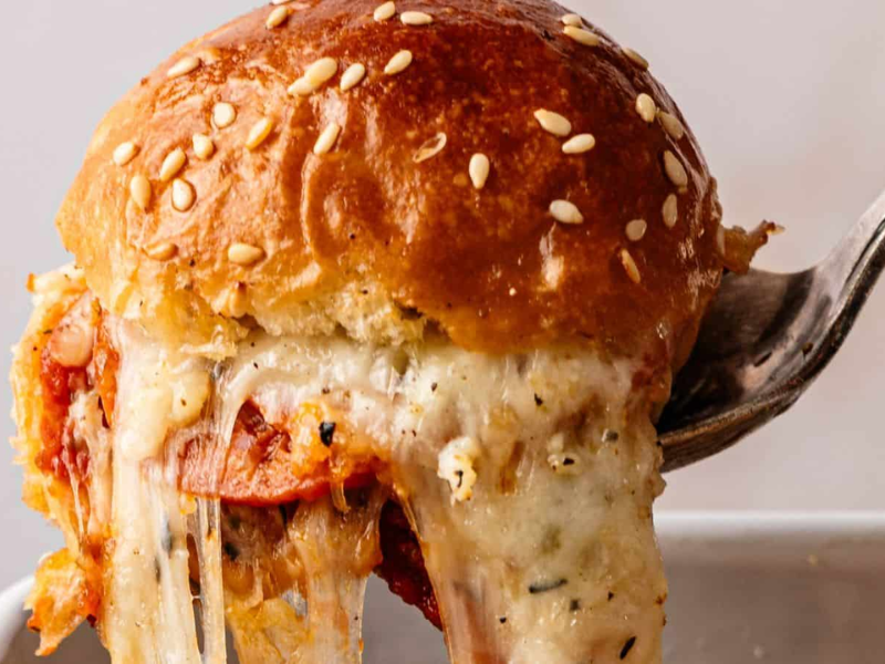 A close-up of a fork lifting a cheesy, sesame-topped slider filled with tomato sauce and melted cheese.