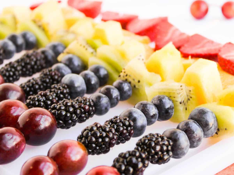 Skewers with grapes, blackberries, blueberries, kiwi, pineapple, and strawberries arranged in neat rows on a white surface.