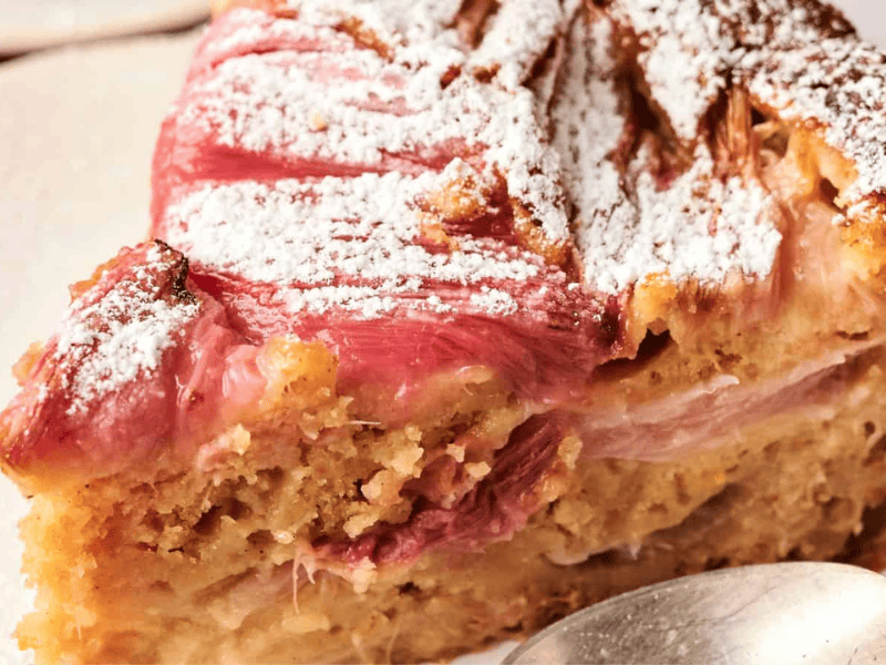 Slice of rhubarb cake dusted with powdered sugar, on a white plate, with a silver spoon beside it.