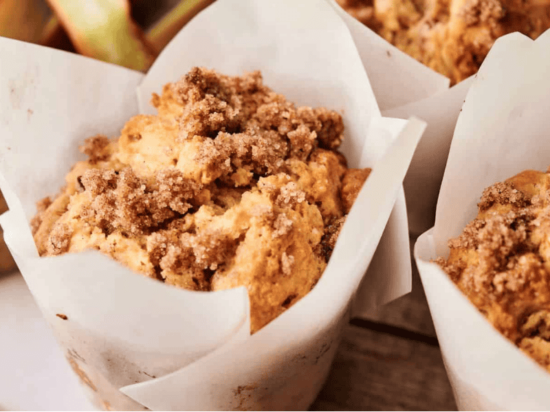 Close-up of three muffins with crumbly streusel topping, each wrapped in white parchment paper, arranged on a wooden surface.