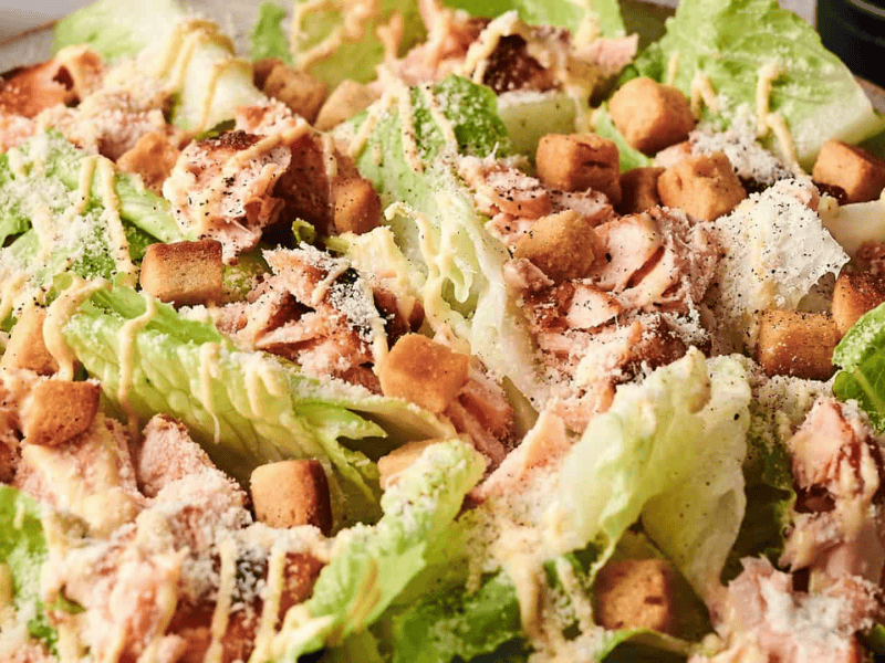 Close-up of a Caesar salad with romaine lettuce, croutons, shredded chicken, grated cheese, and a creamy dressing.