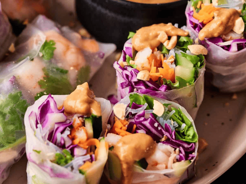 Fresh spring rolls filled with shrimp, purple cabbage, carrots, greens, and topped with chopped peanuts and peanut sauce, served on a plate.