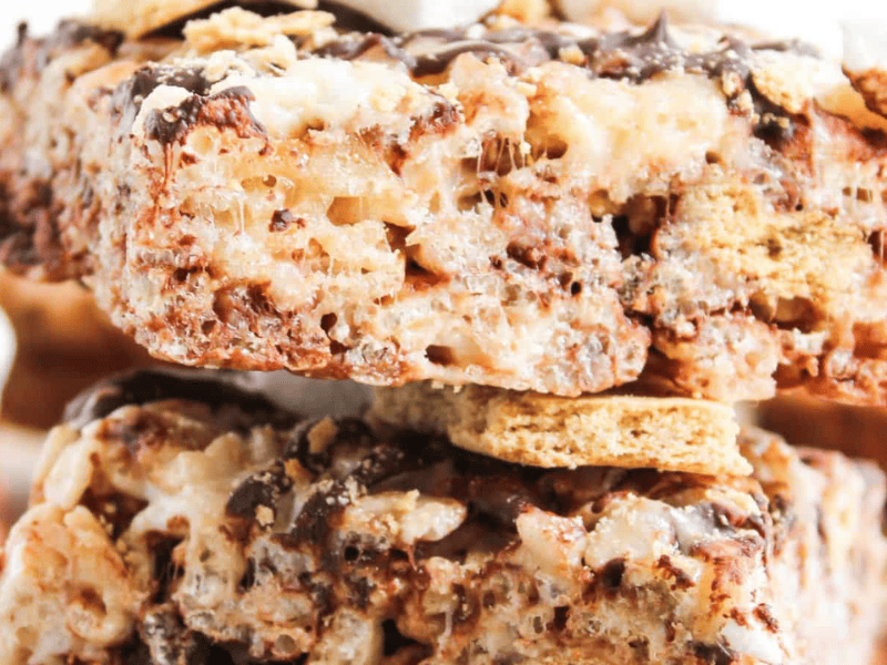 Close-up of stacked dessert bars made with crispy rice cereal, marshmallows, graham cracker pieces, and chocolate drizzle.