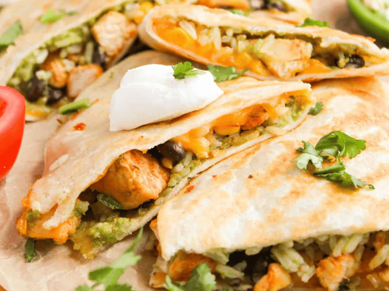 Quesadillas filled with rice, beans, and chicken, topped with sour cream and cilantro. Sliced tomato and jalapeño are on the side.