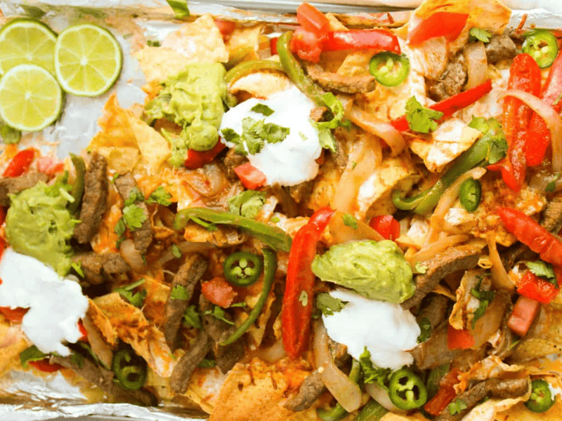 A tray of loaded nachos topped with sliced beef, red and green peppers, jalape&ntilde;os, guacamole, sour cream, and lime wedges on the side.