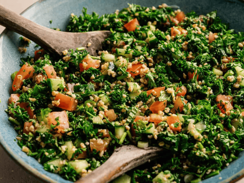 A bowl of tabbouleh salad with chopped parsley, tomatoes, cucumbers, and bulgur, served with wooden utensils.