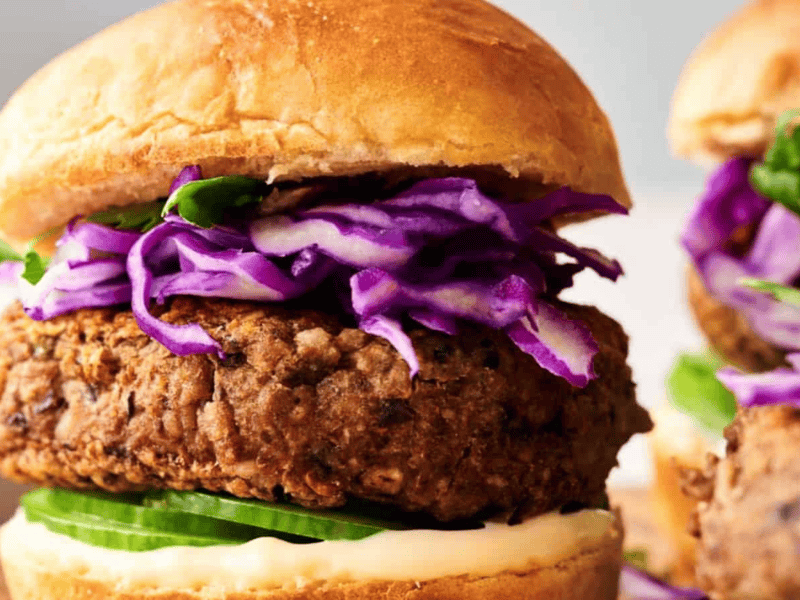 Close-up of a Veggie Burger Slider with a fried patty, sliced cucumber, shredded purple cabbage, greens, and mayonnaise in a bun. In the background, a blurred portion of another slider and a dish are visible.