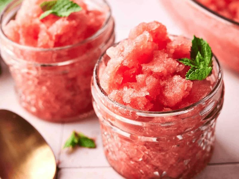 Two small glass jars filled with pink watermelon granita, garnished with mint leaves, are placed on a white tiled surface.
