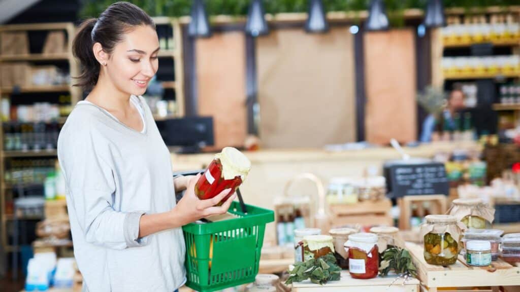 Woman holding a jar of preserved vegetables in a grocery store, examining the label closely&mdash;because food labeling terms aren&rsquo;t simple&mdash;with a green shopping basket in hand.