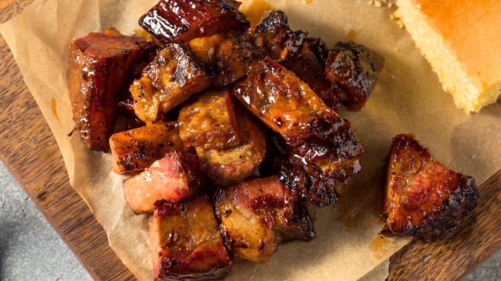 Chunks of barbecued burnt ends on brown parchment paper, served on a wooden surface next to a piece of cornbread.