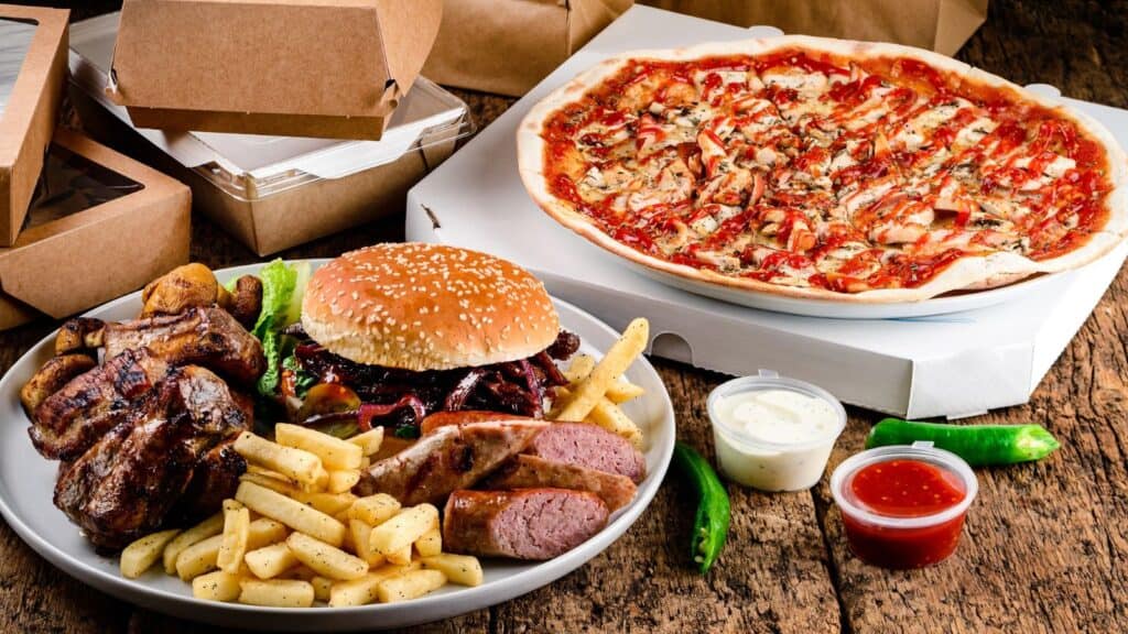 A plate with grilled meats, fries, and a burger&mdash;some of the most-ordered foods in the US&mdash;next to a pizza, dipping sauces, and takeout boxes on a wooden table.