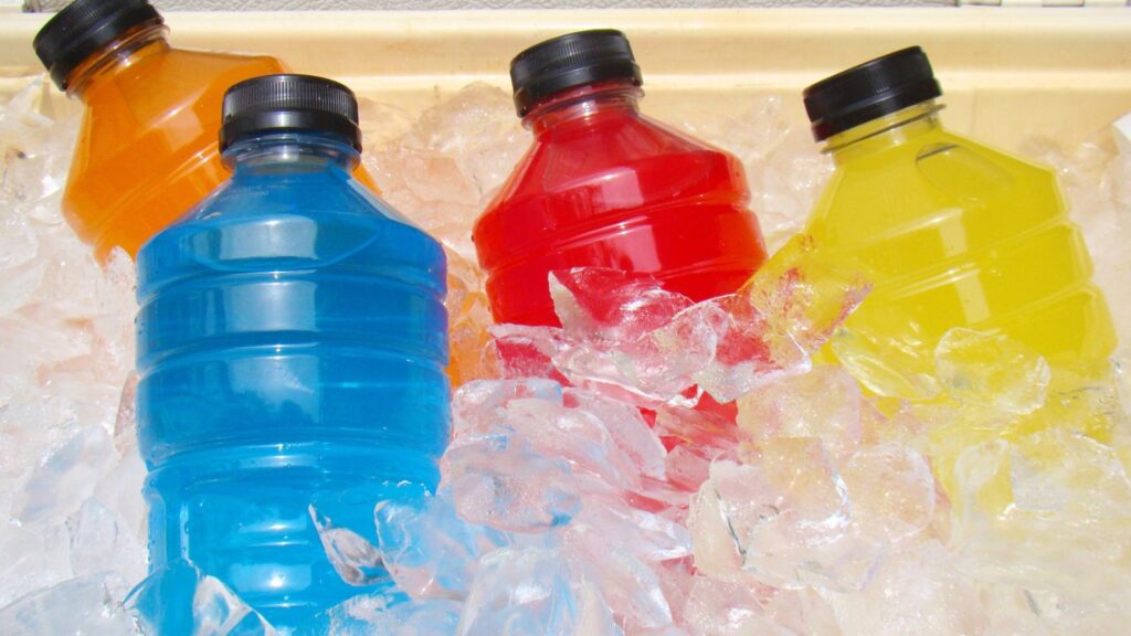 Four plastic bottles with blue, red, yellow, and orange drinks&mdash;healthy foods high in sugar&mdash;are placed on ice cubes inside a cooler. All bottles have black caps.
