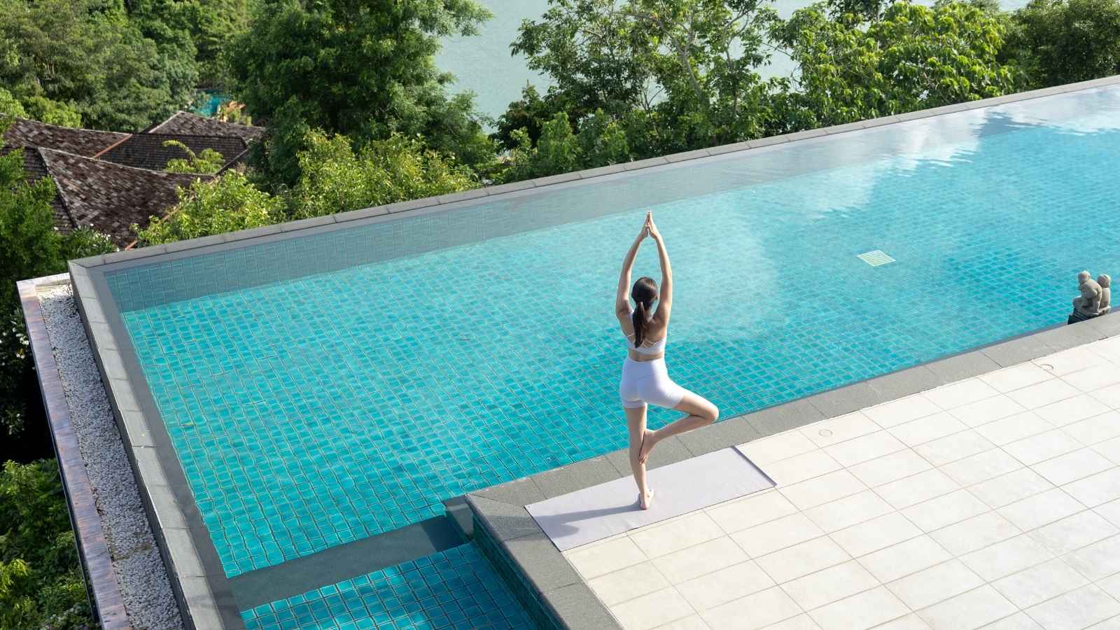A woman practices yoga in tree pose on a mat beside an infinity pool, surrounded by lush greenery and overlooking a body of water.
