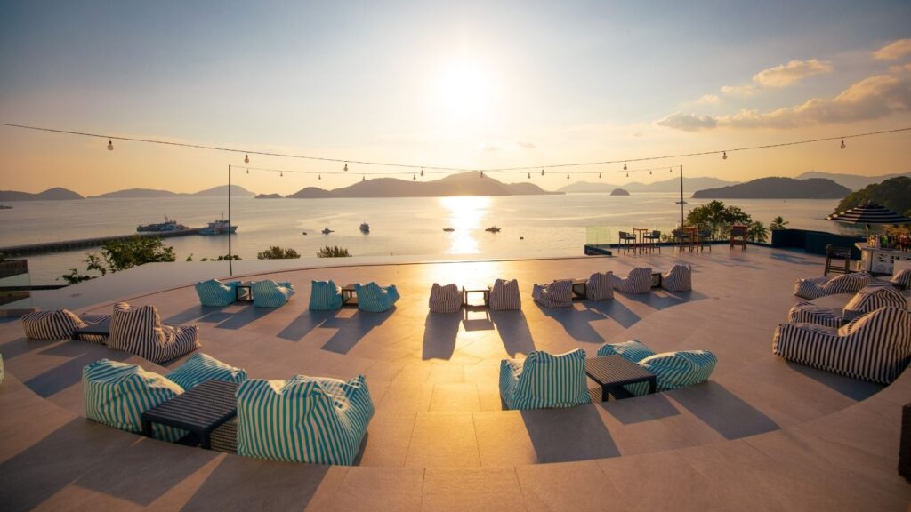 Rooftop terrace with striped bean bags and tables overlooks the sea at sunset, with string lights above and distant islands visible.