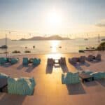 Rooftop terrace with striped bean bags and tables overlooks the sea at sunset, with string lights above and distant islands visible.