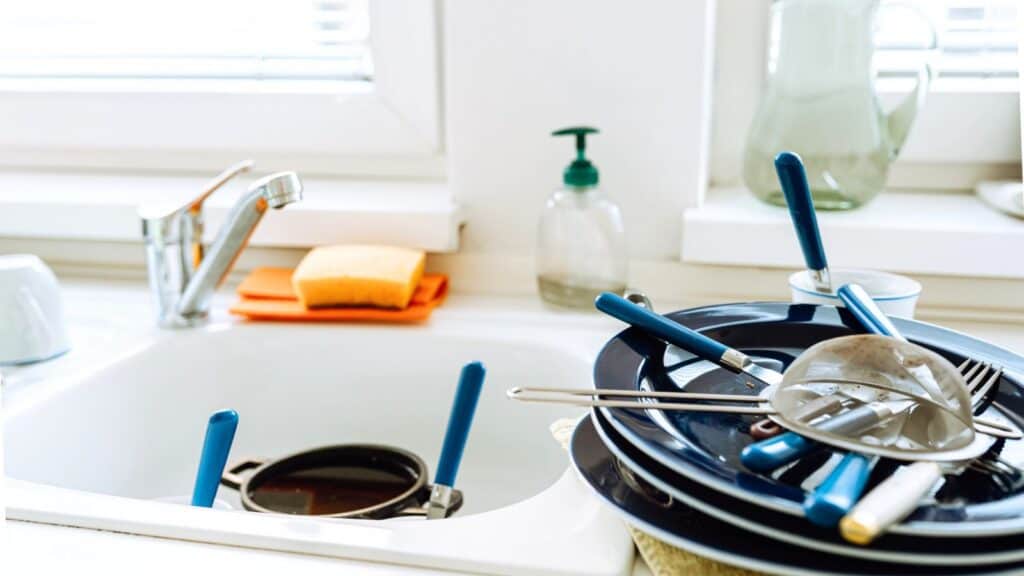 A kitchen sink with dirty dishes, utensils, and pans stacked beside it. A sponge, soap dispenser, and pitcher are on the counter near the window.