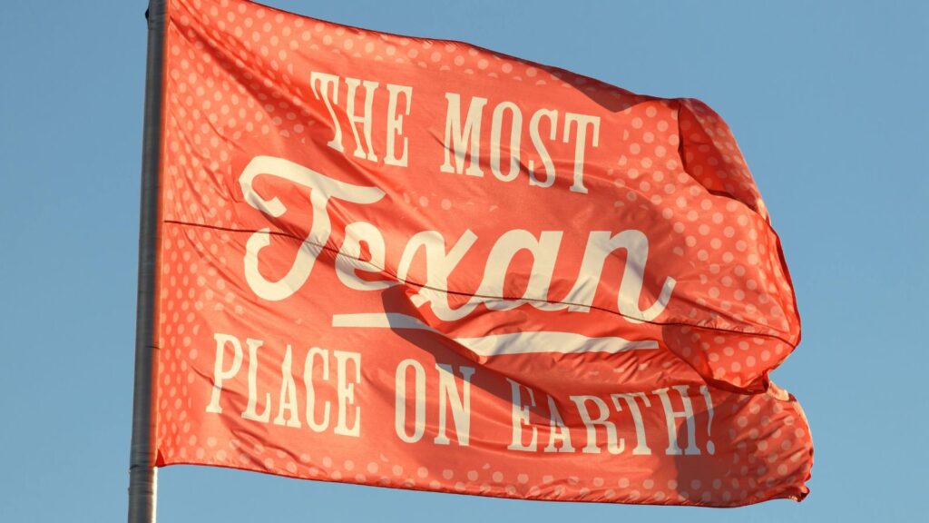 Red flag with white polka dots and text reading &ldquo;The Most Texan Place On Earth!&rdquo;&mdash;a nod to its Texas State Fair finalists status&mdash;waving against a clear blue sky.