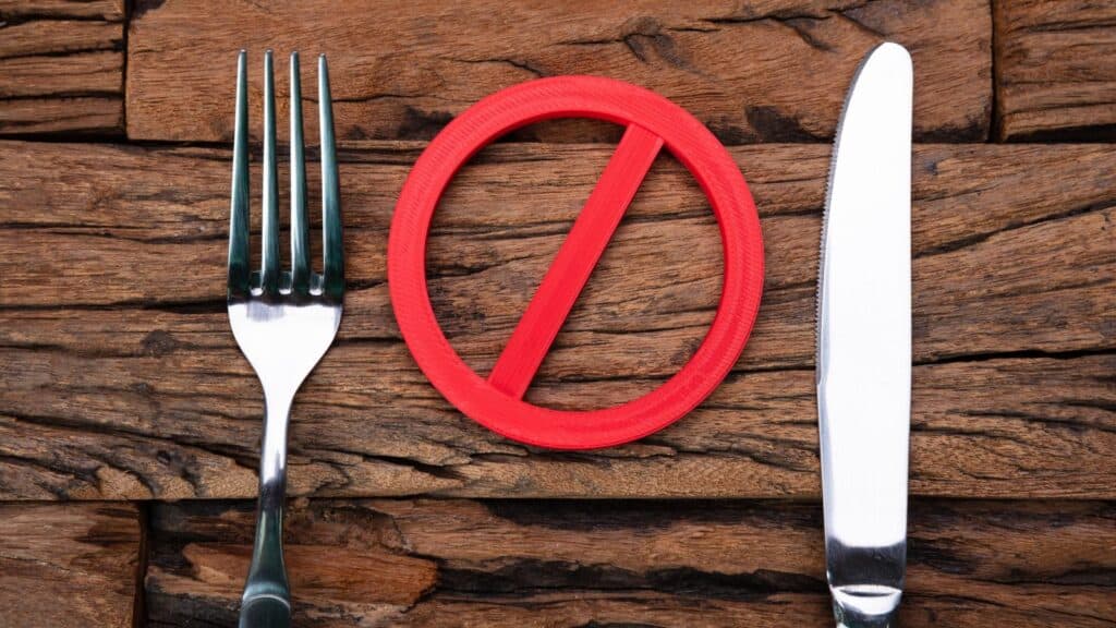 A fork and knife are placed on either side of a red prohibition symbol on a wooden surface, representing how certain foods face bans or restrictions.