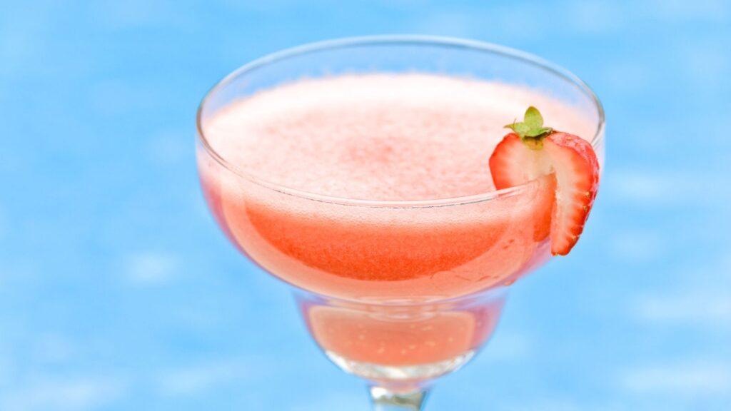 A pink frozen drink served in a glass with a sliced strawberry garnish, set against a blue background.