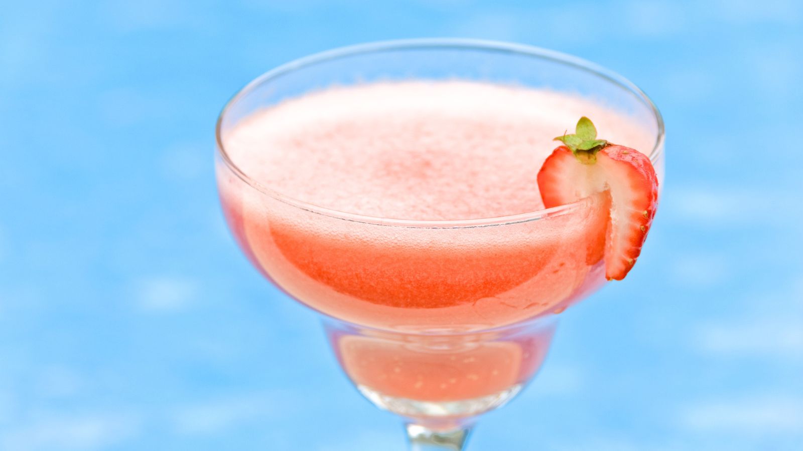 A pink frozen drink served in a glass with a sliced strawberry garnish, set against a blue background.