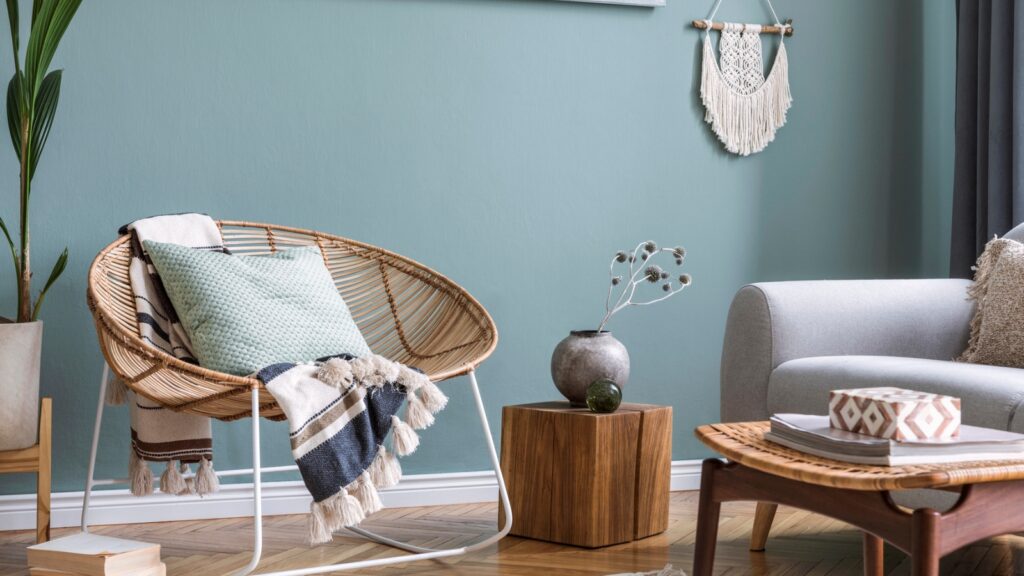 A cozy living room with a rattan chair, green pillow, striped throw, wooden side table, ceramic vase, and a grey couch against a blue wall with macrame art.