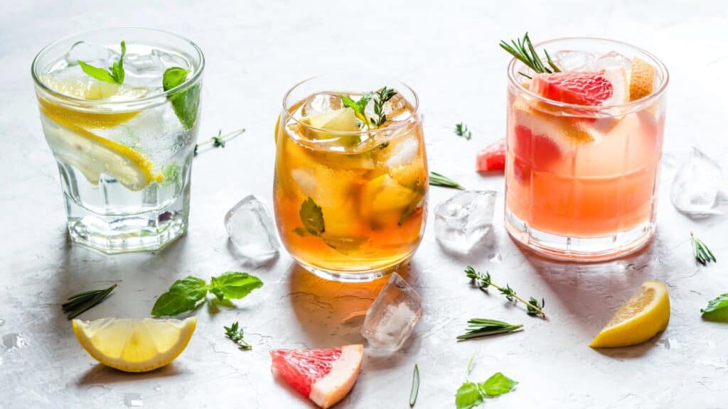 Three glasses of colorful cocktails with ice, garnished with herbs and citrus slices, are arranged on a white surface with scattered ice cubes and fruit pieces, creating a vibrant scene reminiscent of zebra striping patterns in the arrangement.